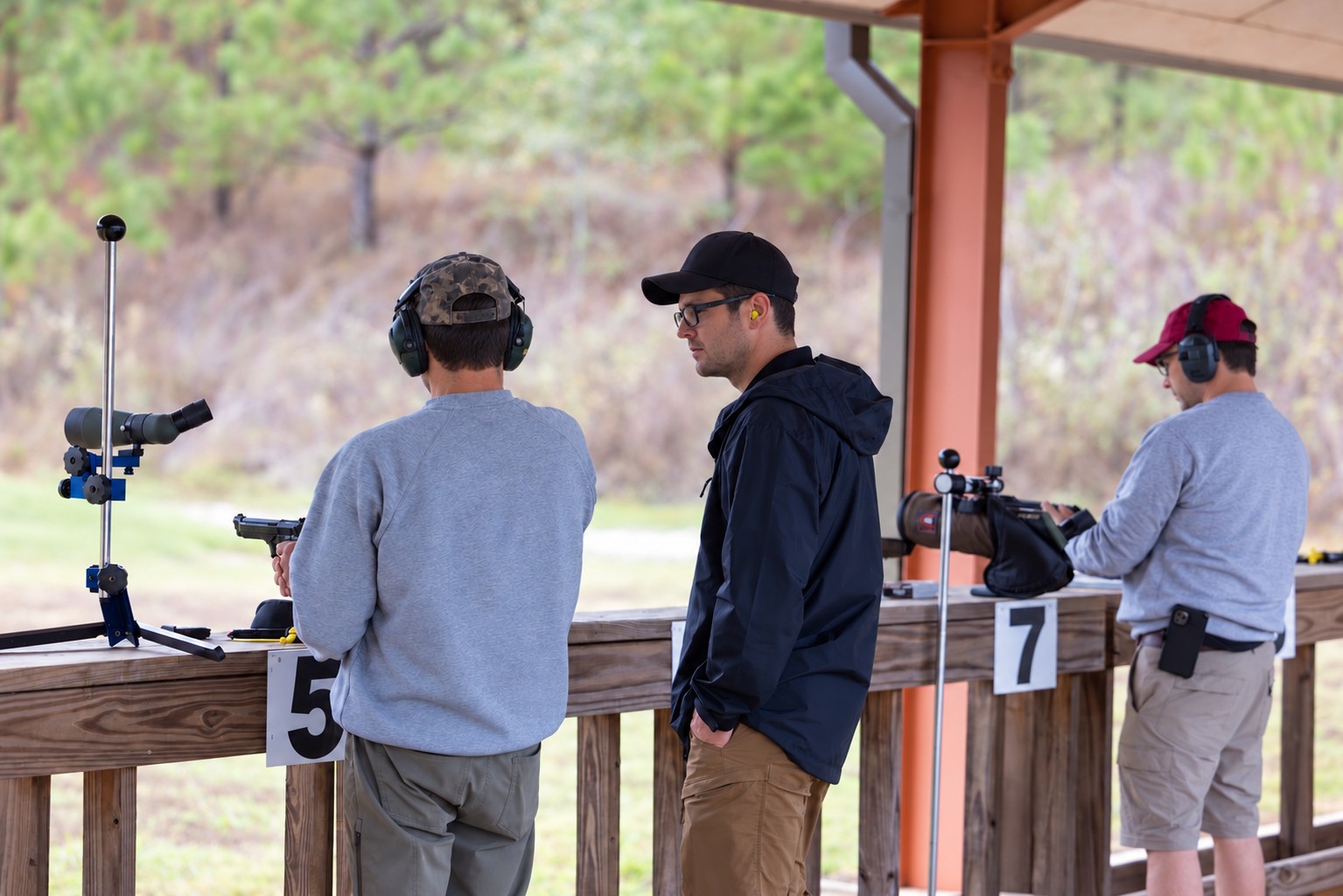 An instructor guiding a student through handgun practice at an outdoor range, both wearing eye and hearing protection.